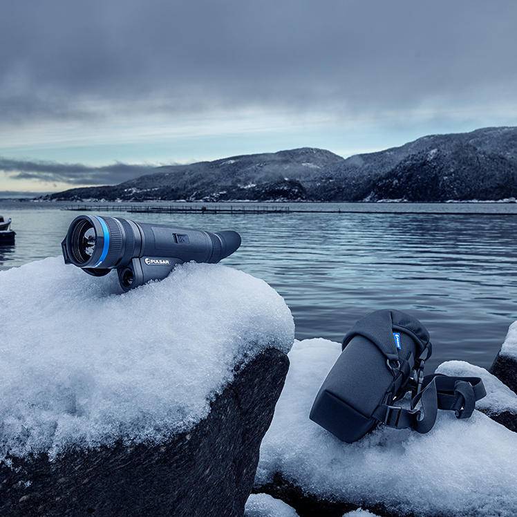Pulsar Telos XP50 thermal monocular placed on snow-covered rock near lake, showcasing portability and outdoor readiness.