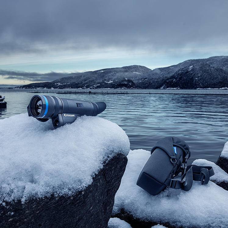 Pulsar Telos XQ35 LRF thermal monocular placed on snow-covered rock with carrying case, highlighting its portability and durability.