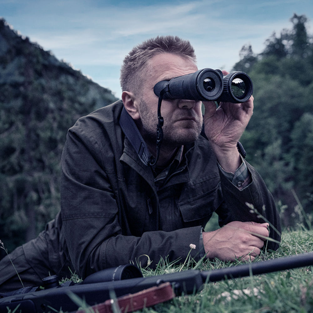 Man using Pulsar Merger XP35 LRF thermal binoculars while lying on grass during a hunt. Perfect for wildlife observation and thermal tracking.