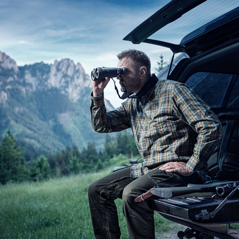 Man sitting on vehicle tailgate using Pulsar Merger XP35 LRF thermal binoculars in mountain terrain. Great for scouting and long-range detection.