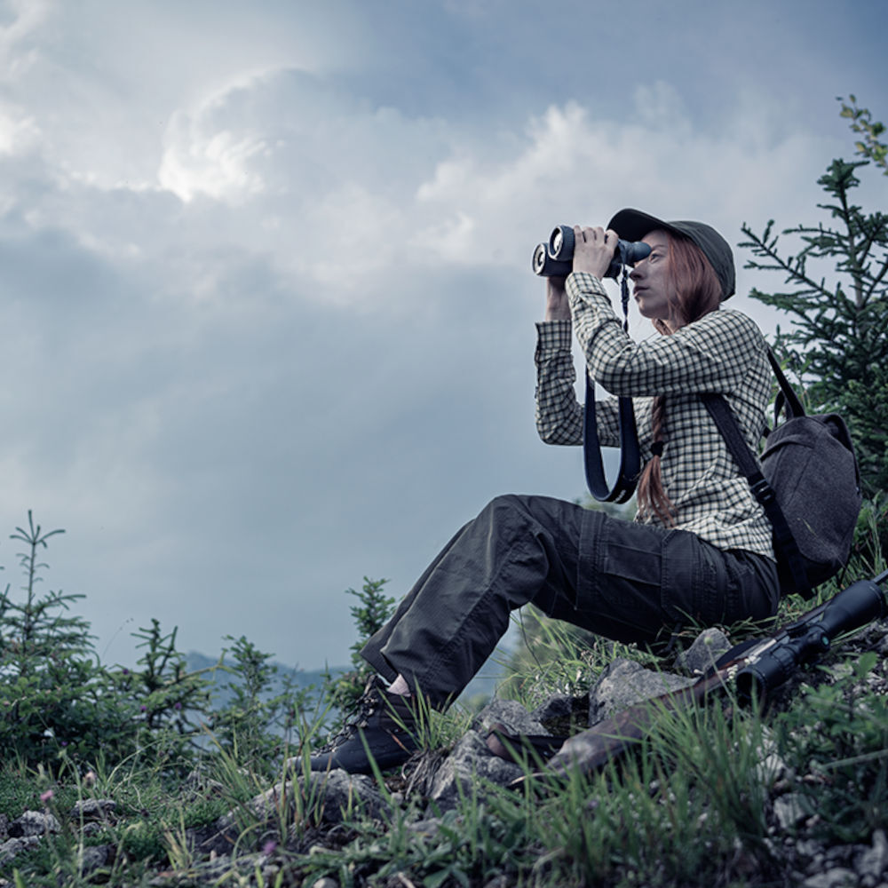 Woman seated on rocky slope using Pulsar Merger XP35 LRF thermal binoculars. Ideal for thermal imaging in rugged outdoor environments.
