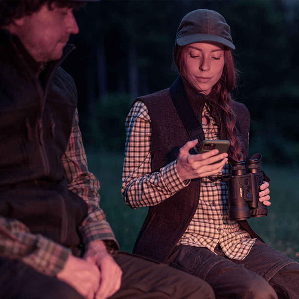 Woman holding Pulsar Merger XP35 LRF thermal binoculars while checking phone near campfire. Combines advanced optics with outdoor convenience.