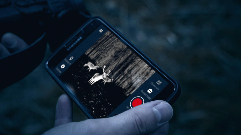 User holding smartphone displaying thermal video of deer via Stream Vision App connected to Pulsar scope in low-light conditions.