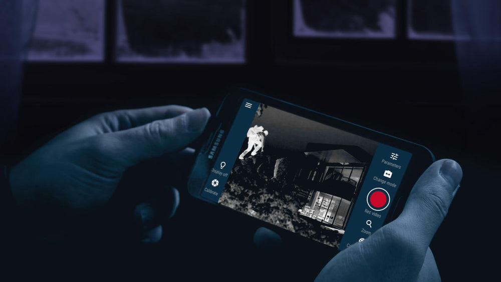 Hands holding smartphone showing thermal image of person near house using Stream Vision App with Pulsar scope for security.